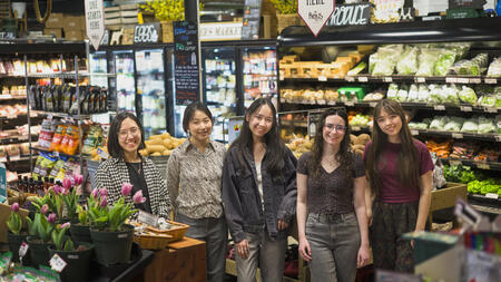Five students pose in a brightly lit produce section of a grocery store