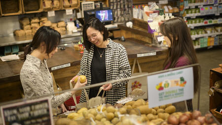 Three students stand beside an onion and potato display, with one selecting food to place into a tote bag