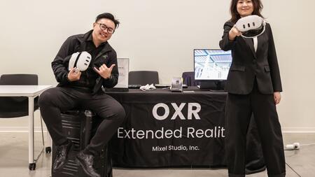 Two people proudly hold headsets in front of a table that reads OXR Extended Reality