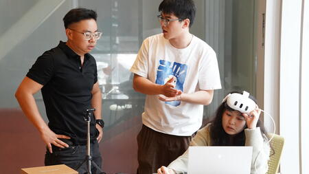 Anhua focuses on a laptop while wearing a headset, with Chiao Lin and another team member standing up and looking on