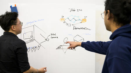 Drawings appear on a white board. Chiao Lin looks at them while Vivek Selveraj points at the white board.