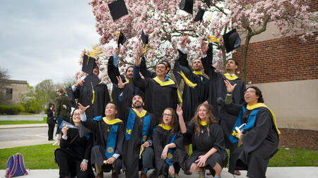 A group of graduates in gowns, tossing their caps in front of a pink flowering tree