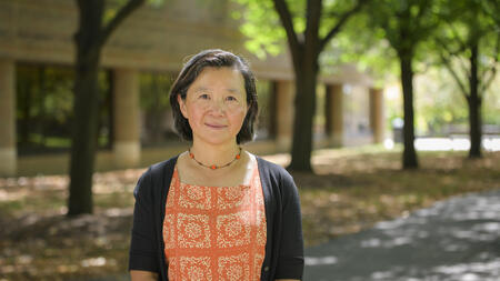 Yan Chen stands outdoors on a tree-lined walkway, wearing an orange floral-patterned dress and a black cardigan.