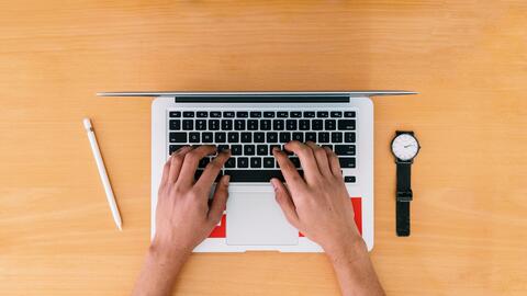 Hand on a laptop keyboard with a pencil and watch on a desk