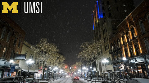 A virtual Zoom background shows a winter scene of Main Street in downtown Ann Arbor on a quiet, snowy night. The upper portion of the First National Bank building is bathed in maize and blue light, and trees wrapped in holiday lighting twinkle on either sides of the street. Upper righthand corner of the virtual background features the UMSI logo. 
