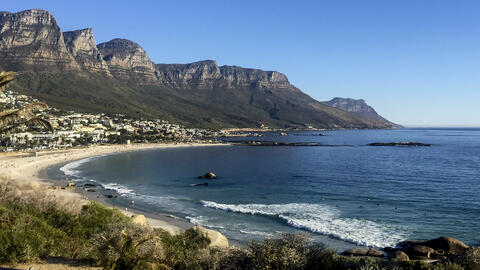 Landscape photo of Cape Town, South Africa coastline. The image captures people on the shore and in the water, and buildings below jagged cliff sides. 