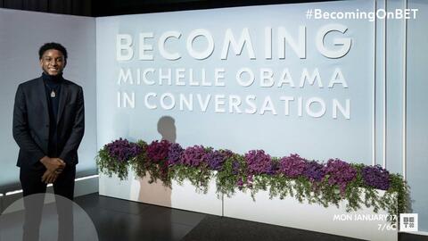 Photo of Joshua Mollison at November 9, 2021 event BECOMING: Michelle Obama in Conversation. He stands and smiles in front of a large sign bearing the title of the event. In front of the sign are two large flower boxes, with leaves and purple flowers spilling over the top. “#BecomingOnBET. Mon. January 17. 7/6C.” BET logo. 