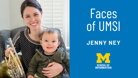 Jenny Ney sits on an armchair, smiling while holding a baby and a French horn. 