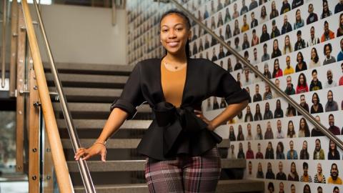 Morgan Miller stands and smiles on a staircase in the Trotter Multicultural Center, in front of a wall decorated with headshots of hundreds of U-M community members. 