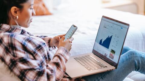 A person lounges with a laptop open on their lap. The screen shows a graph, which they compare against information on their smartphone. 
