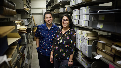 Ricky Punzalan, associate professor of information, and Deirdre de la Cruz, associate professor of history and Asian languages and cultures, stand in the University Library Special Collection between rows of boxes, books and folders of archives. 