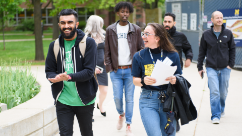 A pair of people smiling and laughing walk among plants and trees outside a University of Michigan campus building. One of them wears a School of Information lanyard and a T-shirt that says “Information changes everything,” and they hold a packet of papers.