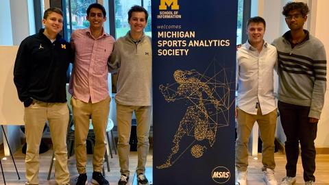 The Michigan Sports Analytics Society leadership board poses with a banner that reads “School of Information welcomes Michigan Sports Analytics Society” at the Michigan Sports Analytics Symposium, March 30, 2022 in North Quad Space 2435. 