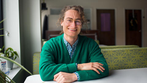 Phil wearing a green cardigan, sitting at a desk next to a plant and smiling