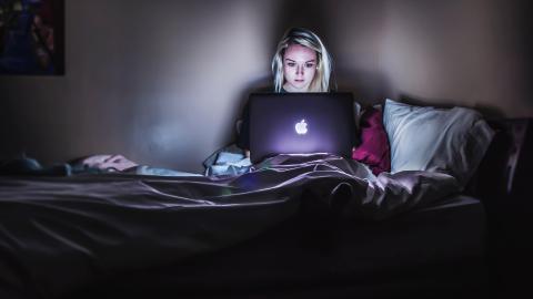 A person sits up in bed and uses a laptop in a dark room.