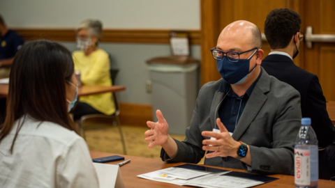 Alumnus Rodger Devine sits across the desk from a current UMSI student. He is masked but speaking animatedly. 