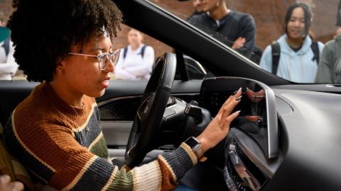A student sits in the drivers seat of the 2023 Cadillac Lyriq and plays with the display of the display. She wears a sweater and glasses.  