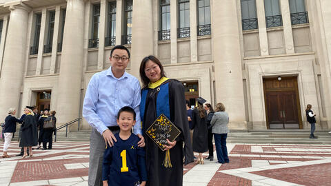Yao Wei dressed in graduation gown stands outside of an auditorium on University of Michigan campus with her husband and son on her graduation day. She holds a decorated graduation cap.