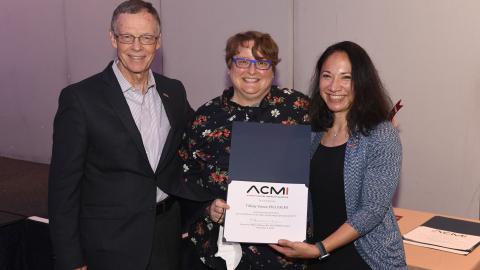 Tiffany Veinot is flanked by the past and present presidents of ACMI. They are smiling and holding an award. 