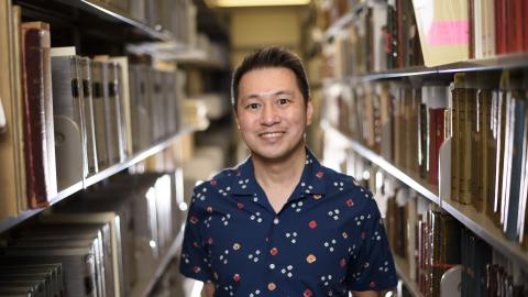 Ricky Punzalan, wearing a blue shirt, smiles as he stands among the stacks of archives in a University of Michigan library. 