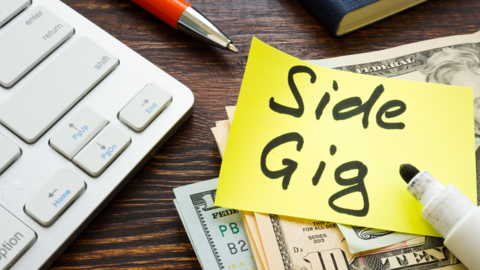 A photo of a keyboard, $10 bills, a pen and a book with a sticky note that says "side gig" in marker. 