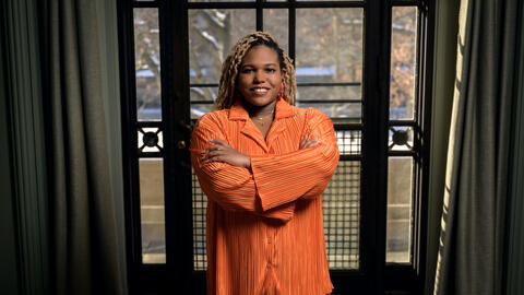 Chelsea Peterson-Salahuddin wearing orange, smiling and standing in front of a window. 