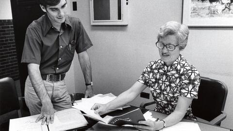 An archival photo of former UMSI professor Constance Rinehart. She is shown sitting at a desk and holding a document. A young man is looking over her shoulder. 