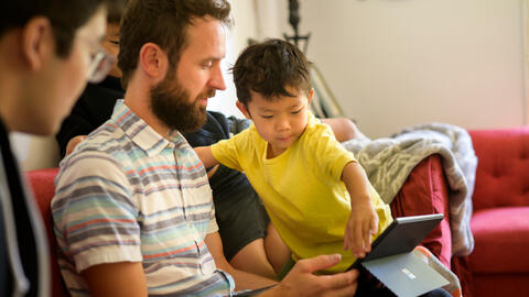 Garret Potter sits on a couch with Max James, holding a laptop. Max reaches toward the laptop screen. Leo looks on from the left side of the frame.