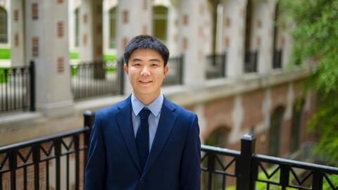Jiaxin Pei wearing a blue suit and tie, standing outside and smiling at the camera. 
