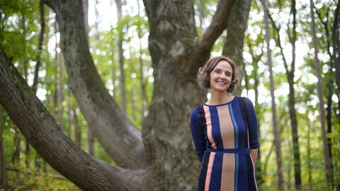 Nicole Ellison in front of a tree on a hiking trail, wearing a blue and pink dress and smiling.  