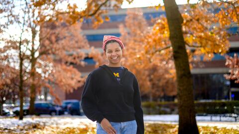 An image of Christine Carethers standing on campus surrounded by fall foliage, wearing a U-M t-shirt and a red bandana