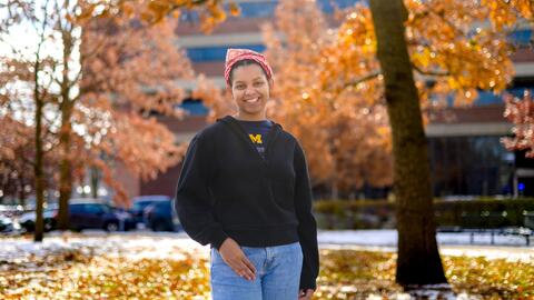 An image of Christine Carethers standing on campus surrounded by fall foliage, wearing a U-M t-shirt and a red bandana