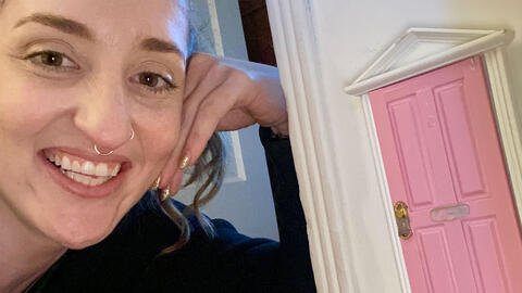 Assistant professor Michaelanne Thomas smiles as she poses next to a pink dollhouse-sized fairy door she installed on the wall of her home. 