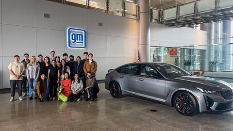 An image of students posing beside a classic Cadillac model in the lobby of General Motors' Cole Building, with an illuminated "gm" sign on the wall behind them