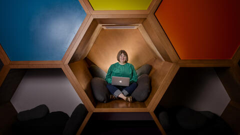 Kristin Fontichiaro sitting cross-legged in colorful cubes at a library, wearing a green sweater and smiling. 