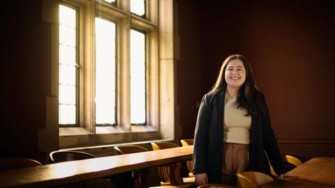 A photo of Julia Kassab posing behind a long wooden desk in an empty Law School classroom