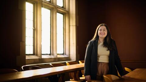 A photo of Julia Kassab posing behind a long wooden desk in an empty Law School classroom