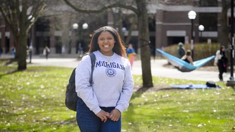 A photo of Ambar Amoros-Gomez standing on the Diag wearing a U-M sweatshirt, with a hammock visible in the background