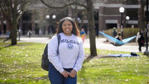 A photo of Ambar Amoros-Gomez standing on the Diag wearing a U-M sweatshirt, with a hammock visible in the background