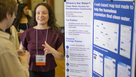 A student stands in front of a trifold poster, chatting animatedly with a guest at Exposition
