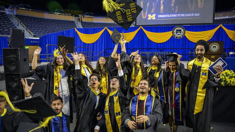 A photo of a group of UMSI graduates cheering in caps and gowns, tossing a cap in the air that reads "Keep calm and go blue"