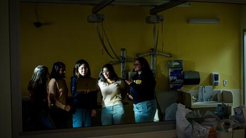 A photo of five students reflected in a mirror in the Clinical Simulation Center, gathered around the glow of a laptop