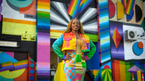 Megan Threats in a colorful jacket and a long skirt standing in front of a multi-colored wall. 
