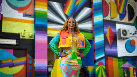 Megan Threats in a colorful jacket and a long skirt standing in front of a multi-colored wall. 