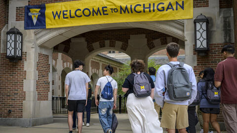 About 10 students walk into an outdoor brick passageway that is decorated with a maize and blue banner that reads "Welcome to Michigan." Two of the students wear UMSI-branded backpacks.