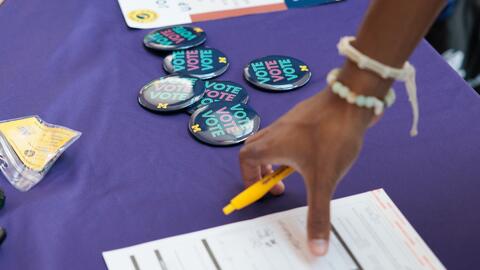 A pile of UMichVotes buttons sit on a table near someone filling out a form.