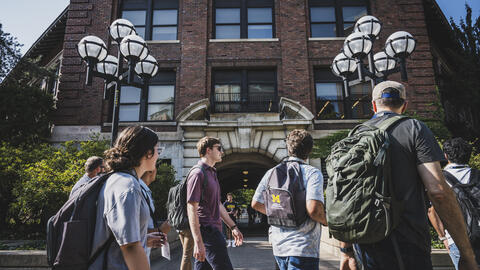 Students wearing backpacks walk on a sidewalk through U-M's Ann Arbor campus. One backpack has a UMSI logo