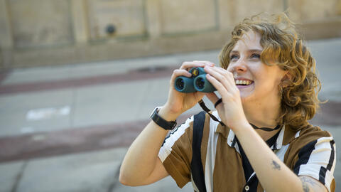 Meg Millure holds a pair of binoculars and smiles while glancing upward 