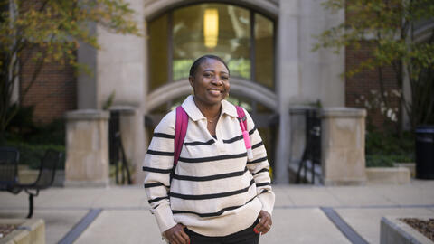 Olga Hamilton smiles in front of the entrance to a campus building, wearing a pink backpack