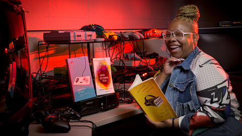 Kishonna sitting in front of a video game, mid-laugh and holding a copy of her book, Woke Gaming. She is surrounded by red light. 